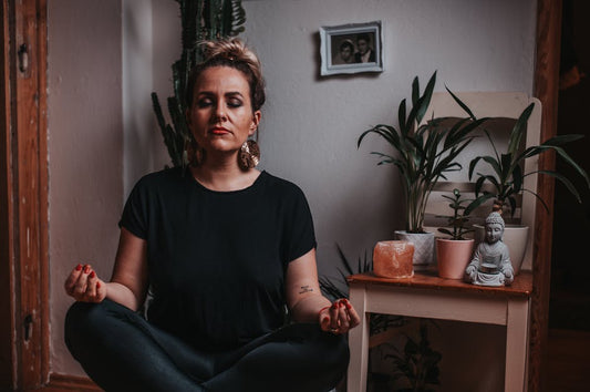 Woman meditating indoors with plants, Himalayan salt lamp, and Buddha statue for wellness