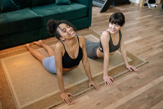 Two women practicing yoga together indoors on a mat, promoting wellness and healthy living.