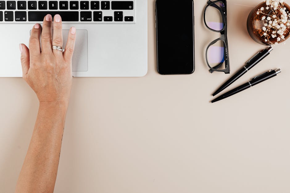 Hand on laptop keyboard with smartphone, glasses, pens, and plant on workspace desk
