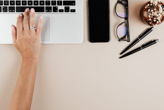 Hand on laptop keyboard with smartphone, glasses, pens, and plant on workspace desk