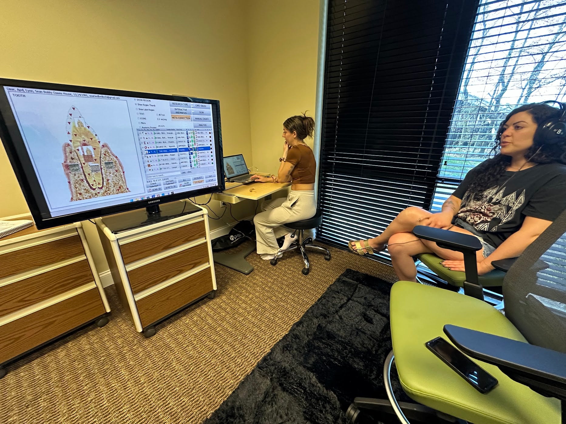 Two people in a room with a large screen displaying a map, one person sitting at a desk with a laptop.