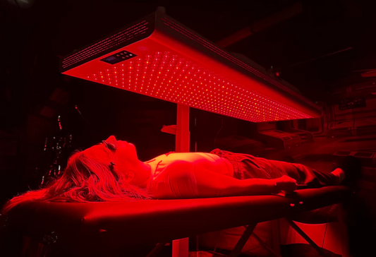 Person receiving red light therapy under a LED panel in a dimly lit room.