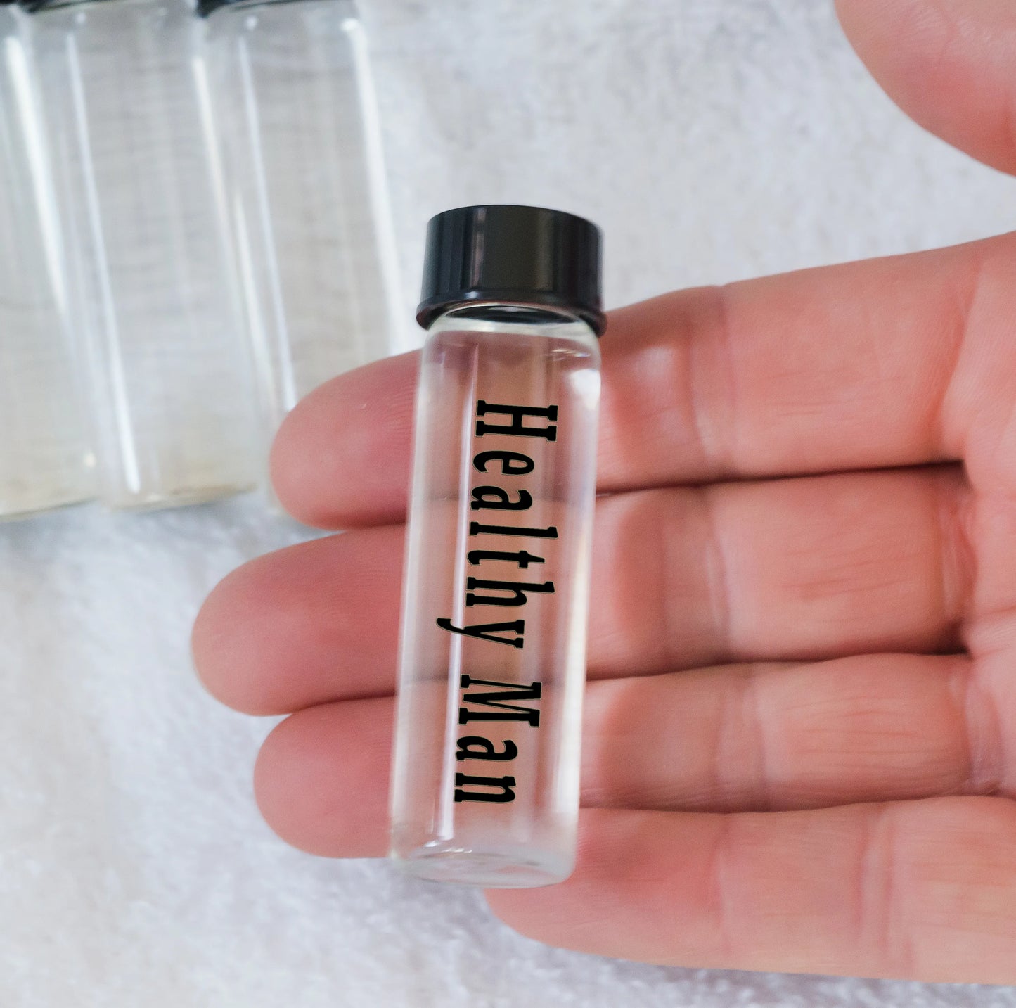 A hand holding a transparent plasma energy water vial labeled 'Healthy Man' with a black cap, in front of a large clear glass or plastic container.