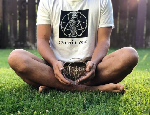 Person sitting on grass holding a small object with 'Omni Core' design on a white t-shirt.