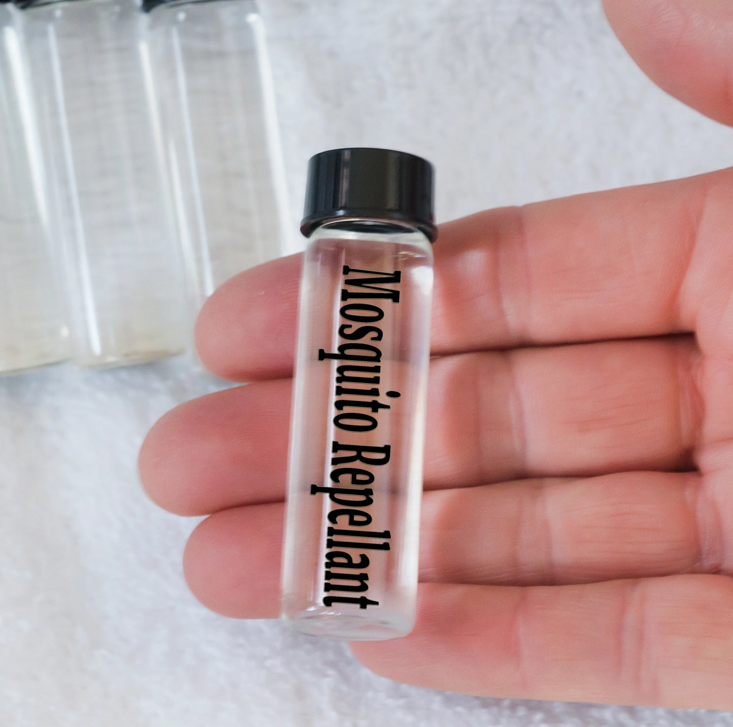 A hand holding a small clear glass bottle labeled 'Mosquito Repellent' with a black cap, filled with a clear liquid. plasma energy vial 